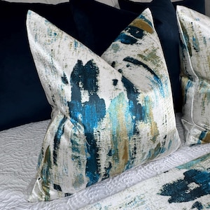 May include: Decorative throw pillows with a white background and a blue, teal, and gold abstract brushstroke design. The pillows are placed on a white quilted bedspread, with navy blue pillows in the background.