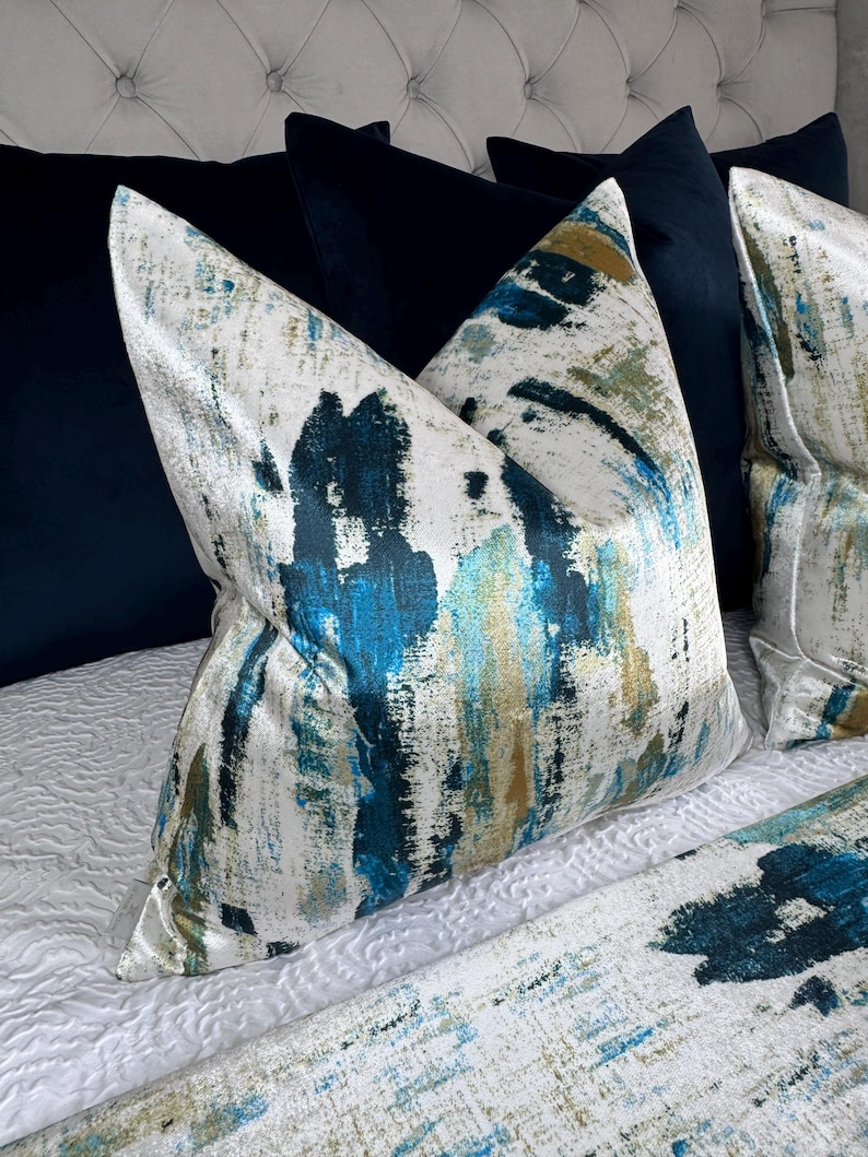 May include: Decorative throw pillows with a white background and a blue, teal, and gold abstract brushstroke design. The pillows are placed on a white quilted bedspread, with navy blue pillows in the background.