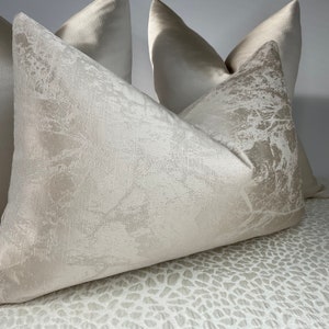 May include: Three decorative throw pillows with a subtle textured pattern in a light beige colour. The pillows are arranged on a white textured surface.