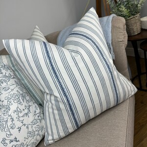 May include: Two decorative throw pillows with a white background and blue stripes. The pillows have a subtle blue stitching detail around the edges.
