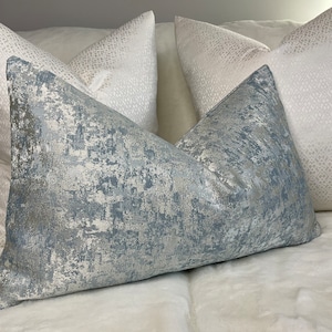 Puede incluir: Tres almohadas decorativas, una con un patrón abstracto azul y plateado, y dos con un patrón geométrico blanco y plateado.