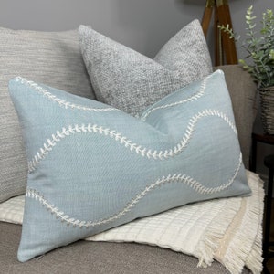 May include: A light blue throw pillow with white embroidered stitching in a wavy pattern. The pillow is on a white throw blanket with fringe.