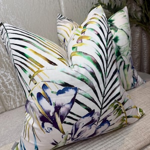 May include: Two square throw pillows featuring a white base fabric with a vibrant botanical print. The design displays large palm leaves in shades of green, yellow, and black, accented by purple and blue floral elements across the textured surface.