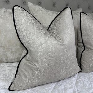 Pode incluir: Close-up de almofadas decorativas com tecido cinza prateado texturizado e debrum preto. As almofadas estão dispostas sobre uma superfície branca estampada, sugerindo um tema de decoração de casa.