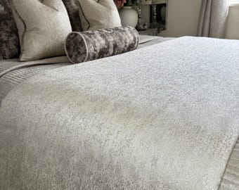 May include: A bed featuring a grey, button-tufted headboard and a selection of decorative pillows. The bed is dressed with a textured, light grey blanket. A cylindrical bolster cushion and square pillows in grey and beige tones are also visible.