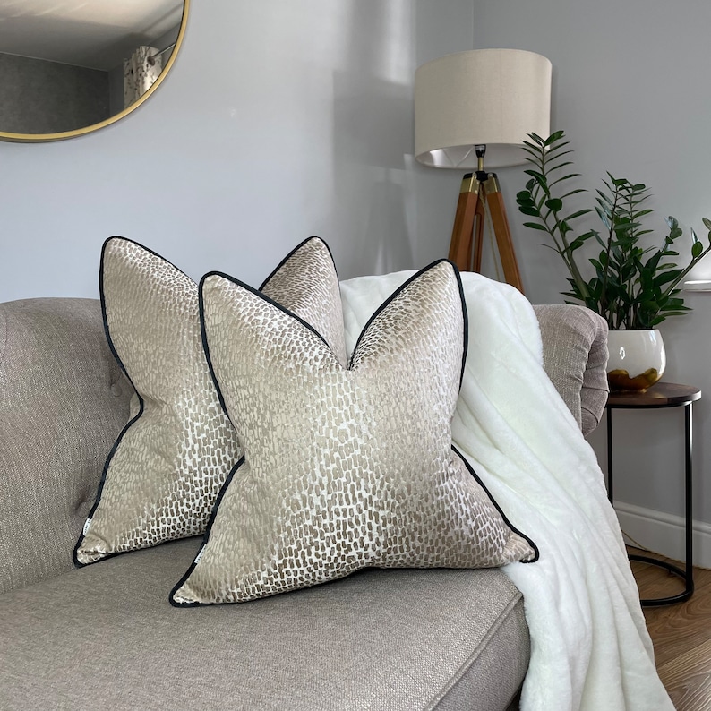 May include: Two beige throw pillows with a black and white abstract pattern and black trim on a gray couch.