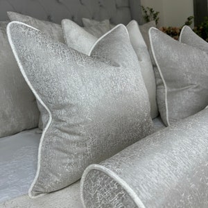 May include: A set of five decorative throw pillows with a silver and white textured fabric. The pillows have a white piping around the edges.