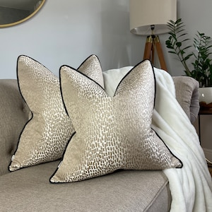 May include: Two decorative throw pillows with a beige and brown abstract pattern. The pillows have black piping around the edges.