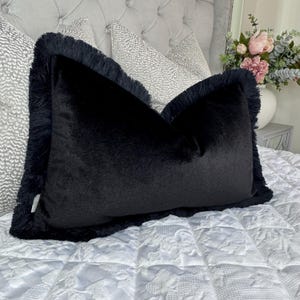 Könnte beinhalten: Ein rechteckiges Kissen aus schwarzem Samt mit Fransenbesatz. Das Kissen ist der Mittelpunkt und liegt auf einer weißen, gemusterten Tagesdecke. Im Hintergrund sind weitere Kissen mit grauen Mustern sowie eine Blumenvase zu sehen.