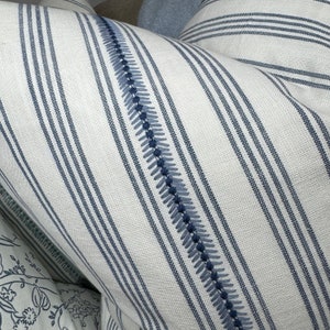 May include: White and blue striped pillow with a blue embroidered detail. The pillow is made of a soft, textured fabric.