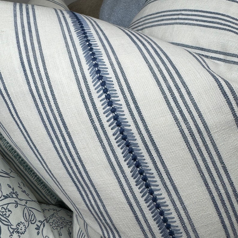 May include: White and blue striped pillow with a blue embroidered detail. The pillow is made of a soft, textured fabric.