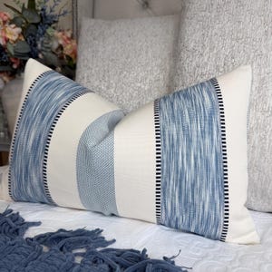 May include: A rectangular decorative pillow with a cream-colored background and blue striped design. The stripes have a textured appearance, with a combination of solid and woven patterns. Black stitching borders the blue stripes.