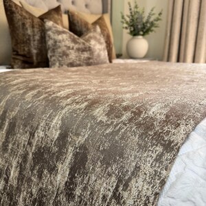 Può includere: Primo piano di un letto con un runner da letto strutturato marrone e beige. Il letto ha diversi cuscini di velluto marrone. Il copriletto ha un aspetto invecchiato e screziato, con una consistenza morbida e lussuosa. Il tema generale è l'arredamento della camera da letto.