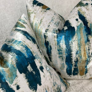 May include: Close-up of two decorative throw pillows. The pillows feature a white background with a bold, abstract design in shades of teal, blue, and gold. The fabric has a slight sheen, adding to the luxurious feel.