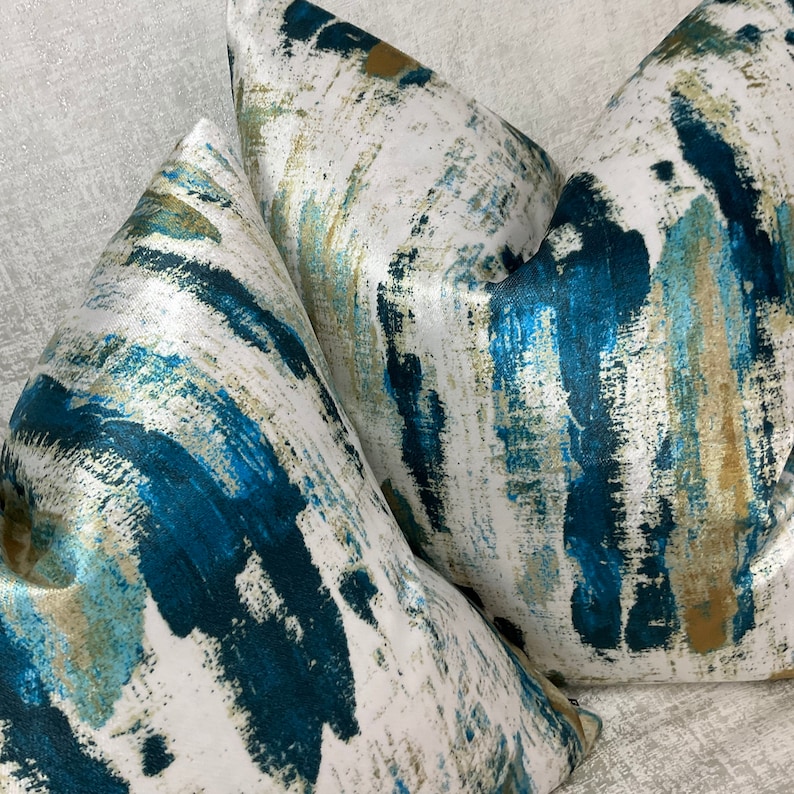 May include: Close-up of two decorative throw pillows. The pillows feature a white background with a bold, abstract design in shades of teal, blue, and gold. The fabric has a slight sheen, adding to the luxurious feel.