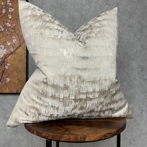 Puede incluir: Una almohada decorativa con un patrón geométrico blanco y dorado. La almohada está sentada en un taburete de madera.