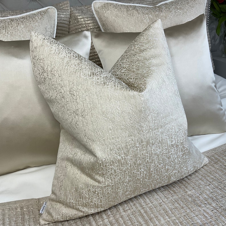 May include: A set of three decorative throw pillows with a textured, shimmery fabric in a light beige color. The pillows are arranged on a bed with a textured beige bedspread.