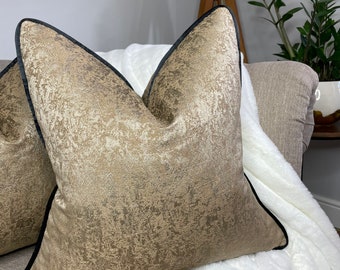 May include: A gold and brown patterned throw pillow with a black trim. The pillow is on a couch with a white throw blanket.