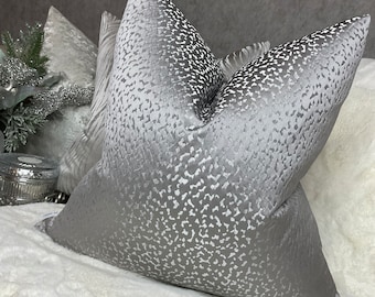 May include: Two silver throw pillows with a white patterned design. The pillows are on a white fluffy surface.