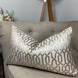 Può includere: Un cuscino decorativo con un motivo geometrico in oro e beige. Il cuscino è su un divano marrone chiaro.