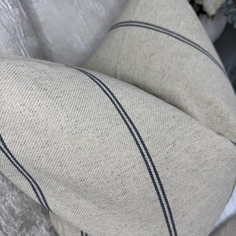 May include: Close-up of two beige throw pillows with dark blue stripes. The pillows are made of a textured fabric and are folded over each other. The pillows are a neutral color and would complement any decor.
