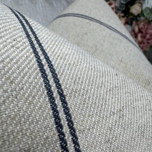 May include: Close-up of a textured, woven fabric in neutral tones. The fabric features two dark blue vertical stripes and a subtle, diagonal weave pattern. The material appears to be linen or a similar natural fiber, with a soft, slightly wrinkled appearance.