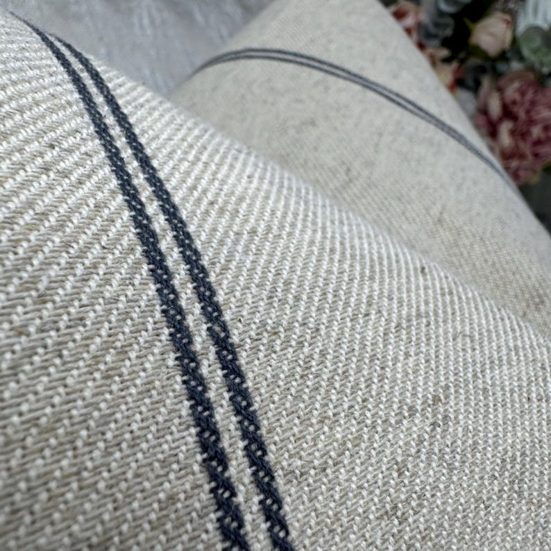 May include: Close-up of a textured, woven fabric in neutral tones. The fabric features two dark blue vertical stripes and a subtle, diagonal weave pattern. The material appears to be linen or a similar natural fiber, with a soft, slightly wrinkled appearance.