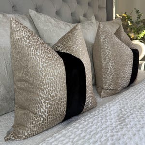 Può includere: Due cuscini decorativi in tessuto beige e marrone con motivo e bordatura in velluto nero. I cuscini sono su un letto bianco.
