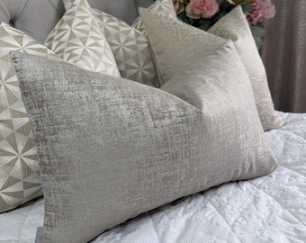 May include: A rectangular throw pillow with a textured, metallic silver-grey finish. It is positioned in front of several other pillows, including some with a geometric starburst pattern in beige and white, and a bouquet of pink flowers.