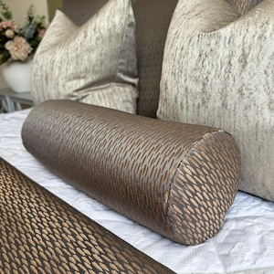 May include: A brown and gold cylindrical cushion with a textured pattern. The cushion is on a white bed with a brown and gold patterned bedspread.