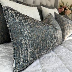 May include: Decorative throw pillows with a textured, abstract pattern in shades of teal, gray, and gold. The pillows are arranged on a white quilted bedspread, with additional white pillows in the background. The pillows add a touch of elegance to the bedroom decor.