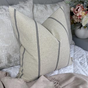 May include: Two beige throw pillows with vertical navy blue stripes. The pillows are on a white quilted bedspread, with a light pink blanket in the foreground. Decorative pillows for a bedroom.