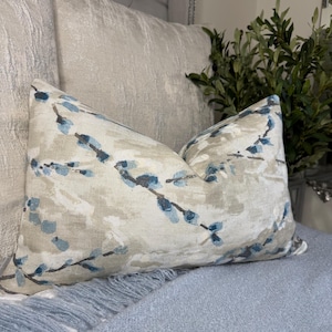Op de afbeelding: Een beige en blauw gepatroneerd kussen met een bloemenmotief. Het kussen ligt op een lichtblauwe deken.