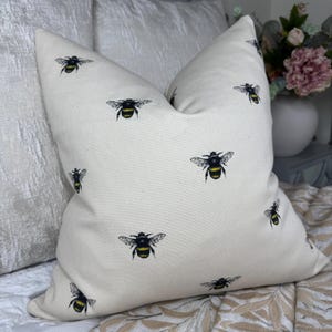 May include: A cream-coloured cushion with a repeating pattern of black and yellow bumblebees. The square cushion is made from a textured fabric. The bees are illustrated with detailed wings and stripes.