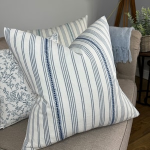 May include: Two decorative throw pillows with a white background and blue stripes. One pillow has a floral pattern in blue and white. The pillows are on a light brown couch.