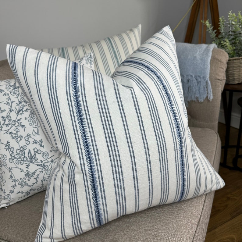 May include: Two decorative throw pillows with a white background and blue stripes. One pillow has a floral pattern in blue and white. The pillows are on a light brown couch.