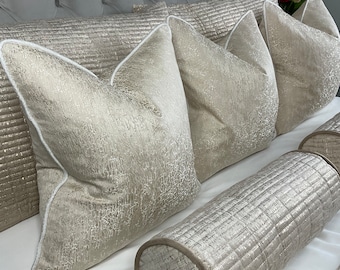 May include: A set of decorative throw pillows and a bolster pillow in a light beige colour with a subtle shimmer. The pillows have a quilted pattern and a white piping trim.