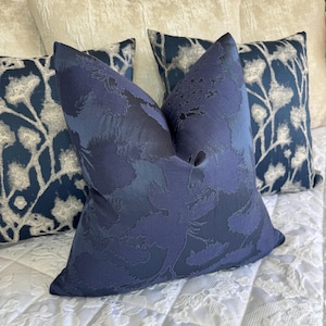 Op de afbeelding: Een verzameling decoratieve sierkussens in tinten blauw en crème. Het voorste kussen is diep marineblauw met een bloemenpatroon. Andere kussens hebben bloemmotieven in zilver en marineblauw. De kussens zijn gerangschikt op een witte, getextureerde ondergrond.