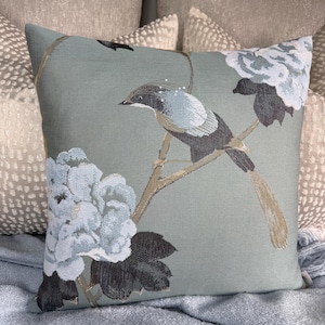 May include: Square decorative pillow with a bird and floral design. The pillow features a light blue background with embroidered details of a bird perched on a branch, and light blue and dark gray flowers.