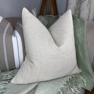 May include: A light beige herringbone pattern pillow is the focal point of this image. A striped pillow and a sage green throw blanket are also visible. The pillows and blanket are on a neutral-toned sofa, creating a cozy, inviting aesthetic.