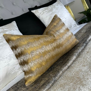 Puede incluir: Una almohada de tiro con estampado dorado y plateado con tejido texturizado. La almohada está sobre una cama con una colcha estampada en blanco y gris.
