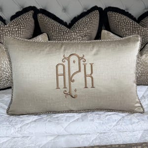 May include: A rectangular beige cushion embroidered with the initials APK in a decorative style. It features a corded trim and is set against a backdrop of spotted pattern cushions with black fringe.