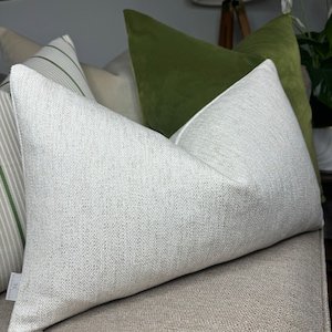 Puede incluir: Una almohada blanca con una superficie texturizada sutil. La almohada está colocada en un sofá beige con una almohada de terciopelo verde detrás.
