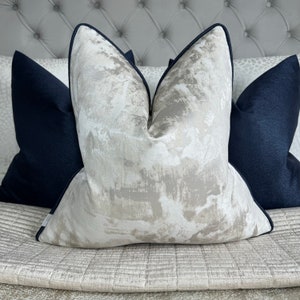 May include: A decorative throw pillow with a white and gold abstract pattern. The pillow has a black trim and is sitting on a grey and white patterned sofa.