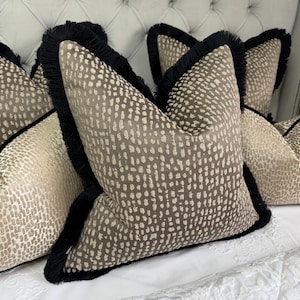 Puede incluir: Cojines decorativos con un estampado moteado marrón y crema. Cada cojín tiene un ribete de flecos negros. Los cojines están colocados sobre una superficie blanca.