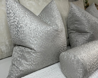 May include: A set of decorative throw pillows with a silver and gray patterned fabric. The pillows are arranged on a bed with a white and gray patterned bedspread.