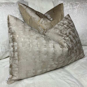 Puede incluir: Tres almohadas decorativas con un patrón geométrico blanco y dorado metálico. Las almohadas están dispuestas sobre una superficie de piel sintética blanca.