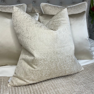 May include: A set of three decorative throw pillows with a textured, shimmery fabric. The pillows are in a light beige color with a subtle sheen. The pillows are arranged on a bed with a beige and white patterned bedspread.