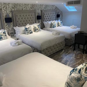 May include: A hotel room with three beds, each with a tufted headboard and white bedding. Decorative pillows with blue and white patterns are on the beds. White towels are neatly arranged on the beds. The room has patterned wallpaper.