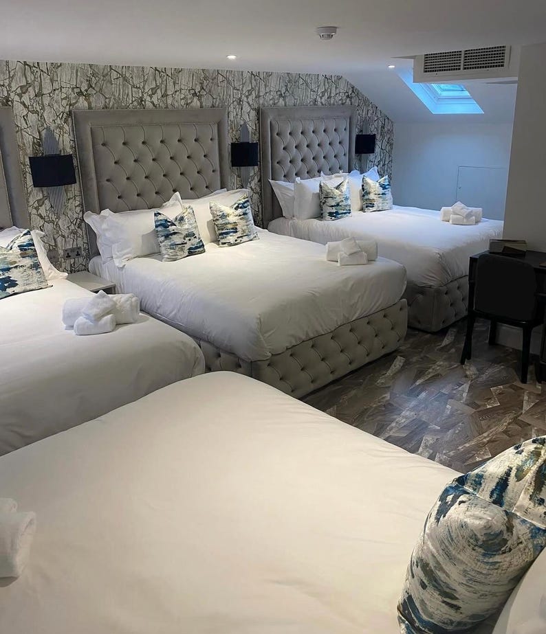May include: A hotel room with three beds, each with a tufted headboard and white bedding. Decorative pillows with blue and white patterns are on the beds. White towels are neatly arranged on the beds. The room has patterned wallpaper.